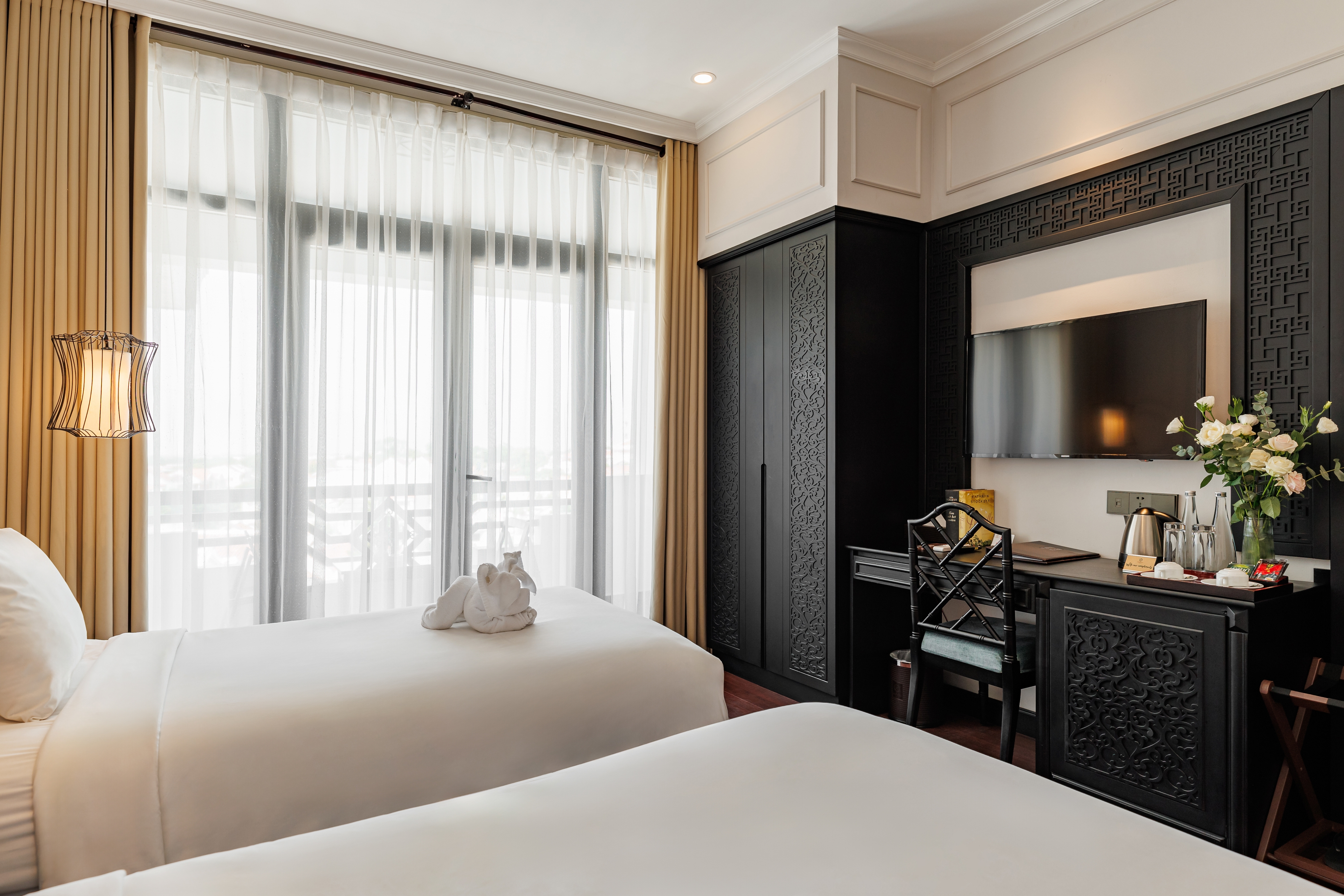 Spacious hotel room with two twin beds, wooden flooring, and large windows adorned with sheer curtains. The room features stylish decor, including a decorative wall piece and a bedside table with a lamp