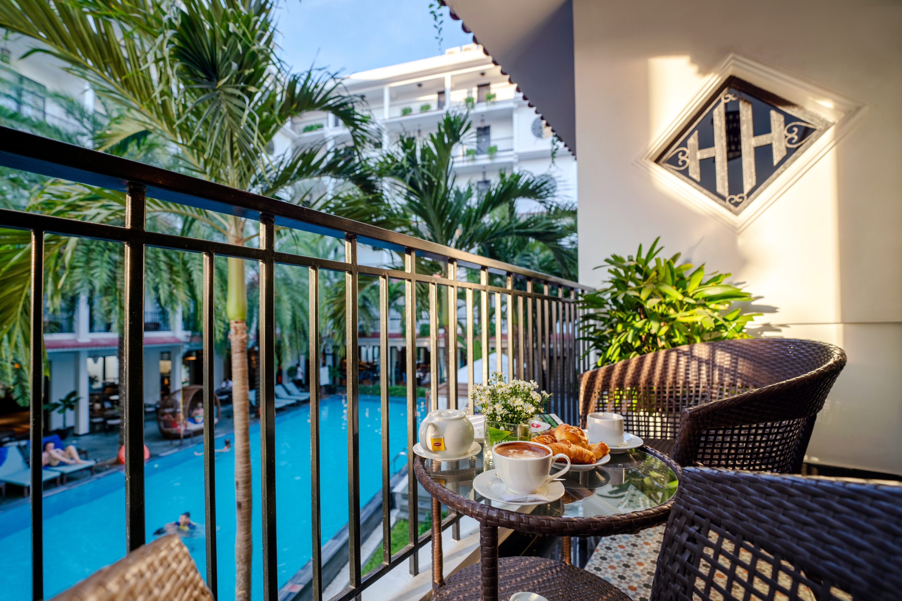 Cozy balcony overlooking a swimming pool, featuring a small table set with breakfast items and surrounded by lush greenery. Sunlight casts shadows on the railing, creating a warm and inviting atmosphere