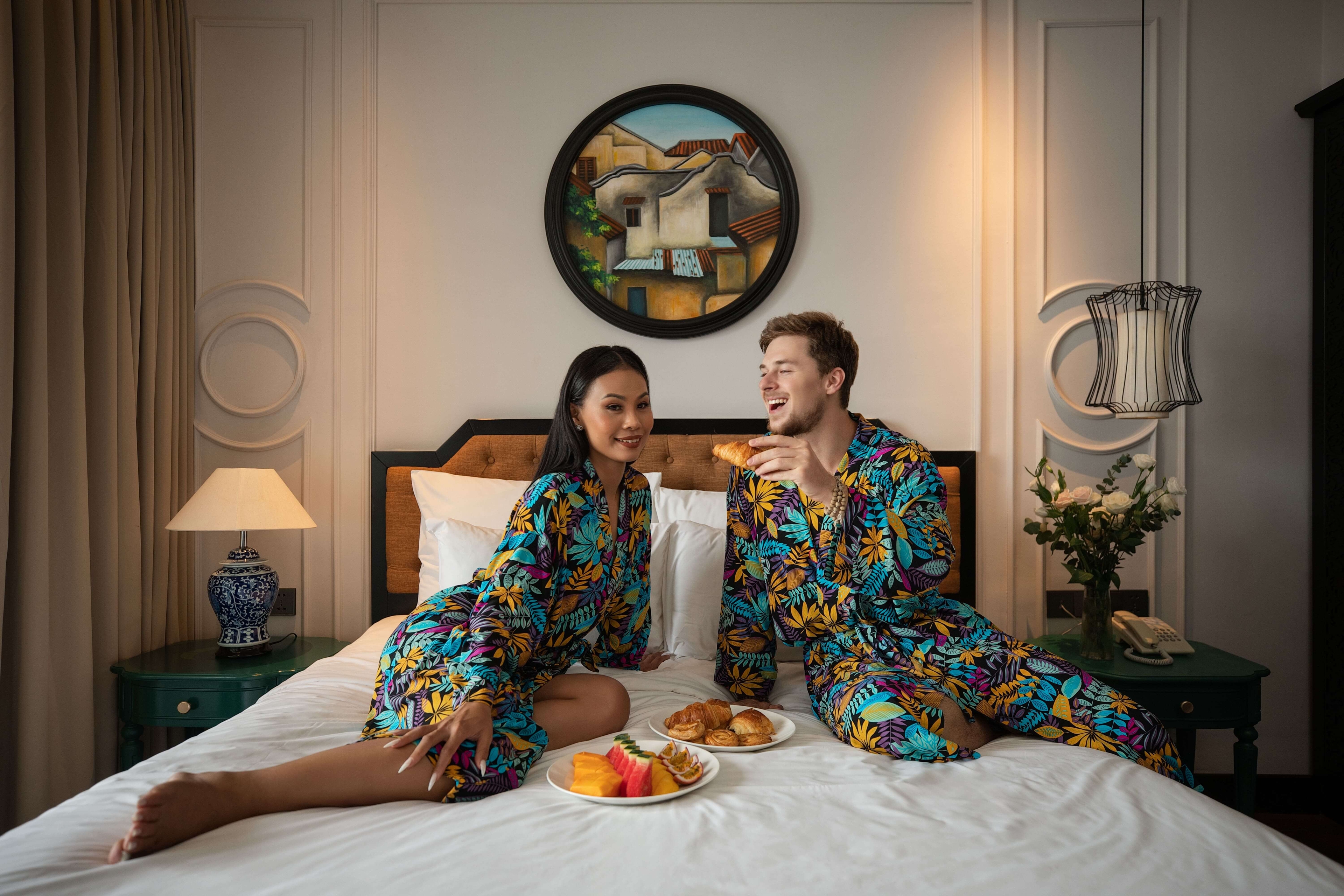 A couple in matching floral robes sitting on a bed, enjoying a breakfast spread that includes fresh fruits and pastries, they appear relaxed and happy