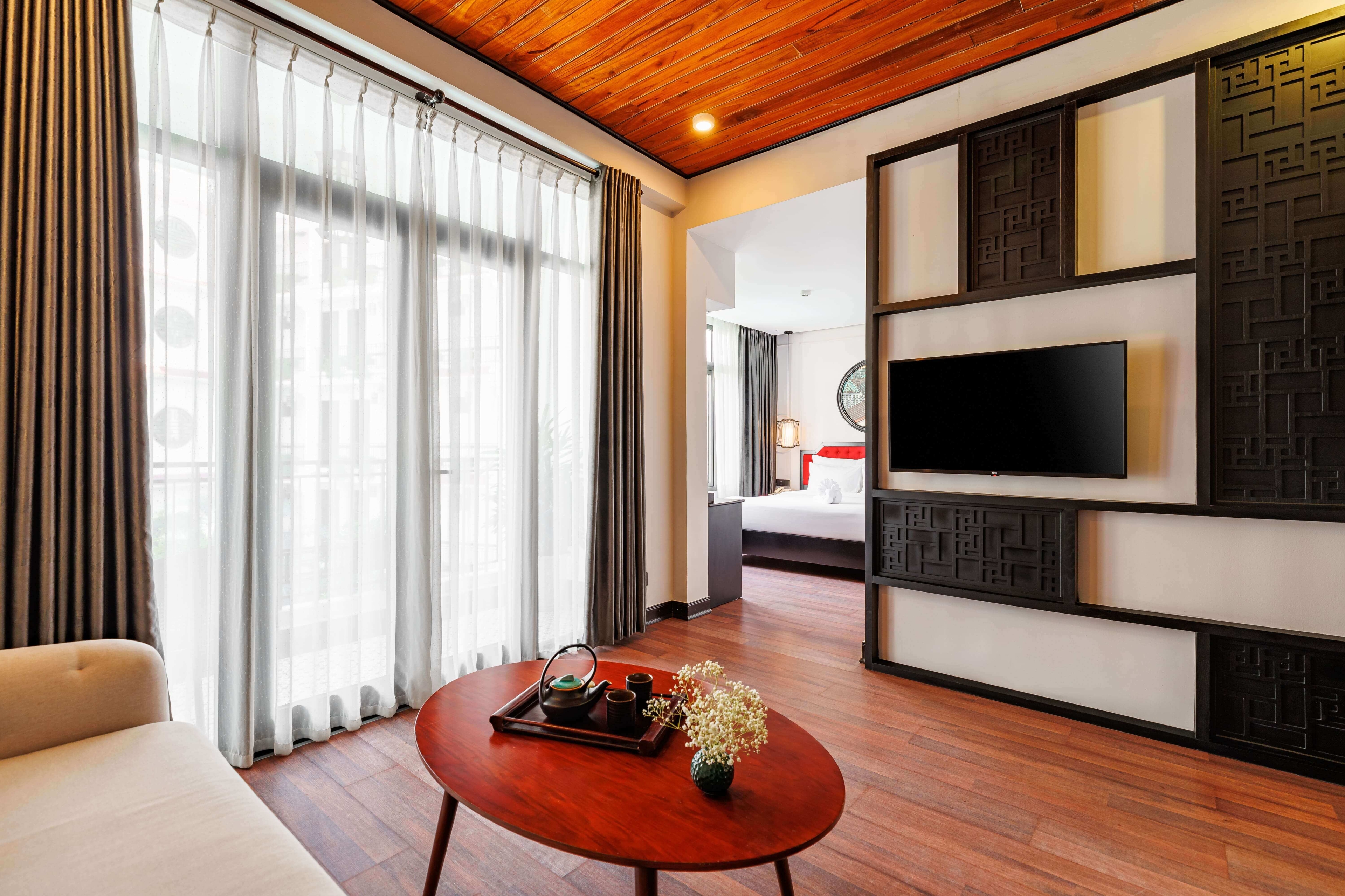 Stylish hotel living area featuring a round wooden coffee table and a comfortable sofa. Large windows with sheer curtains let in natural light, and a modern TV is mounted on a decorative wall unit. The warm wooden ceiling adds a cozy touch to the space