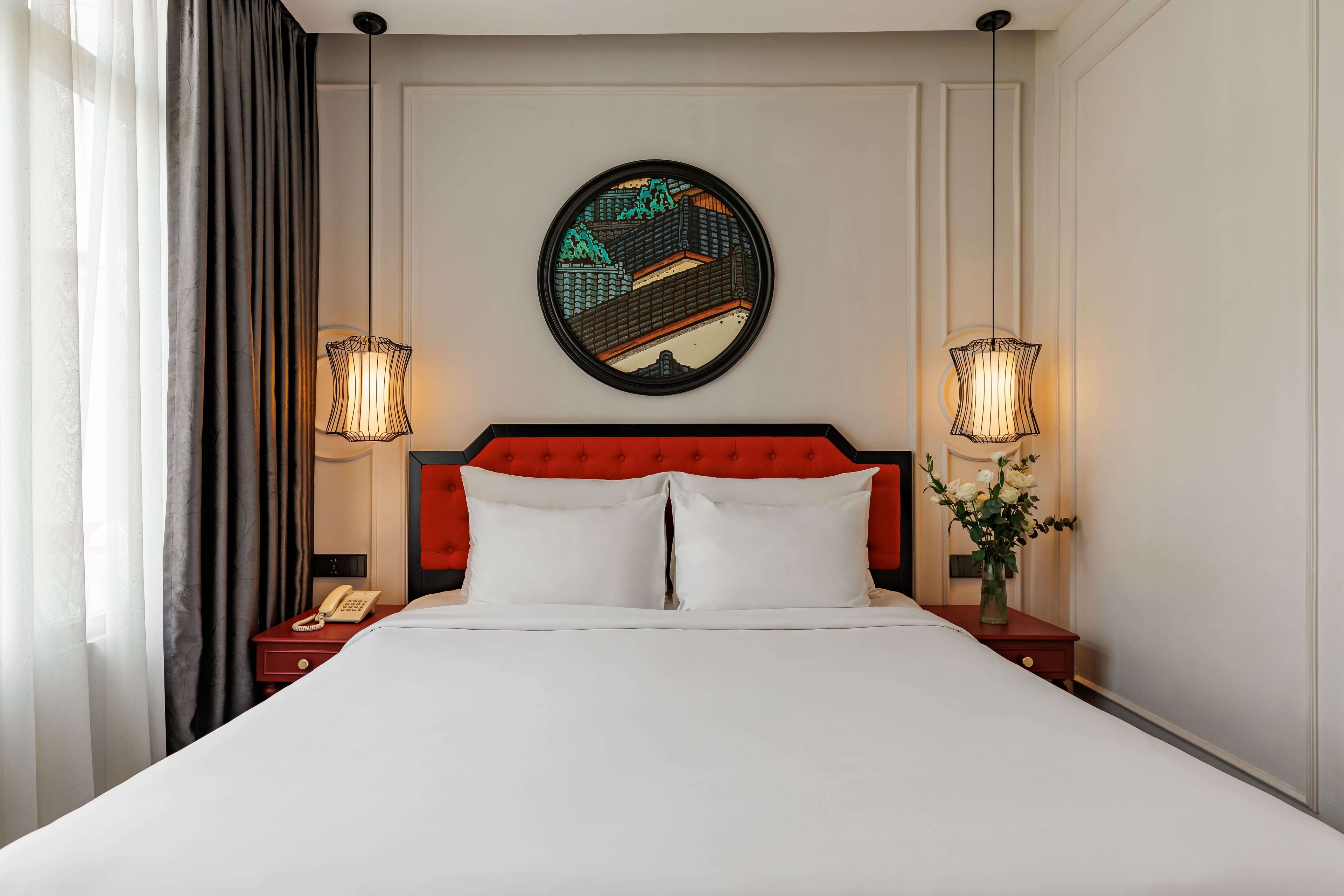 Cozy hotel room featuring a queen-sized bed with crisp white linens and a vibrant red headboard. Flanking the bed are stylish lamps and a decorative round mirror on the wall, complemented by soft natural light filtering through the sheer curtains