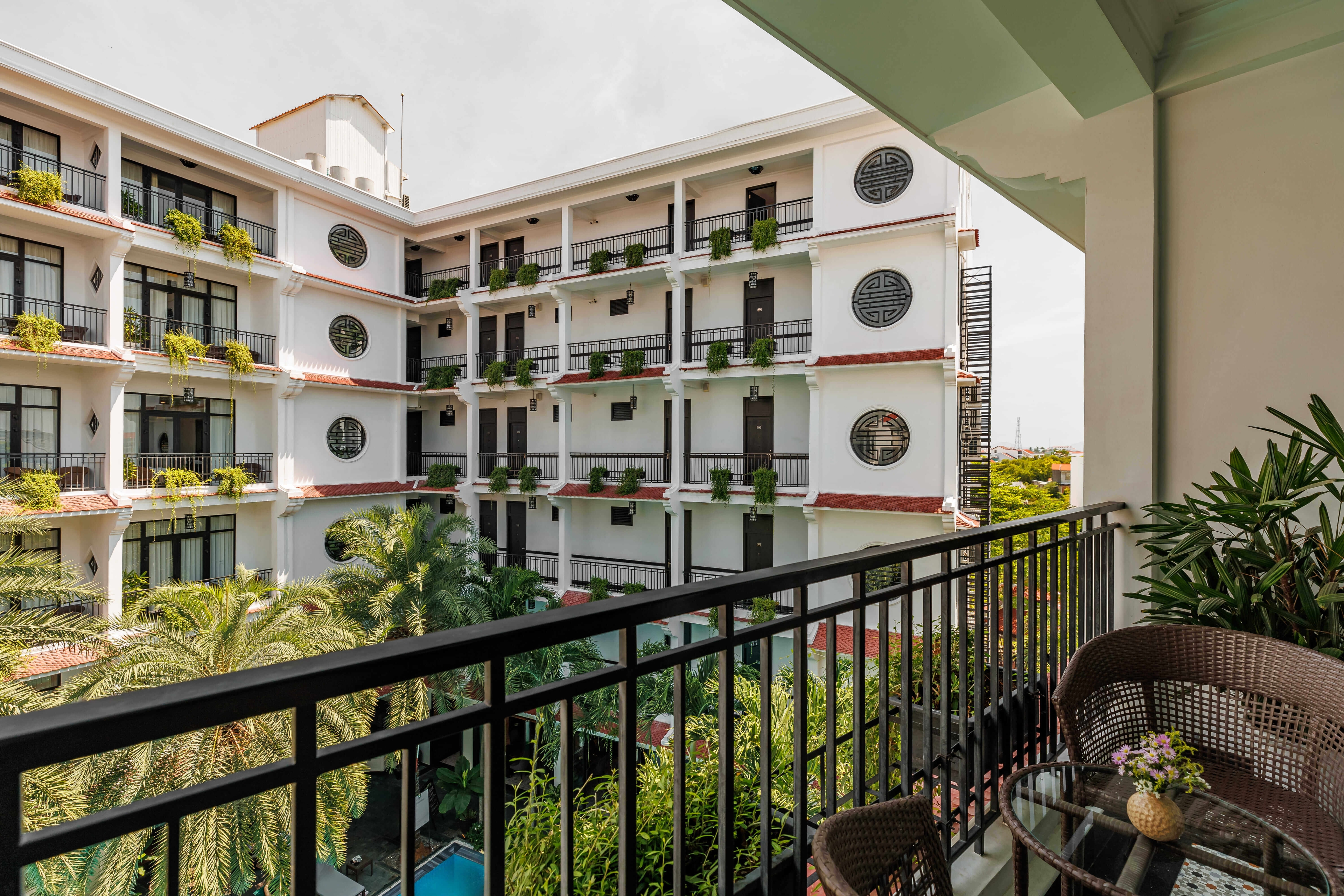 View from a hotel balcony overlooking a lush courtyard with palm trees and a swimming pool. The balcony features a small seating area with two chairs. The surrounding building displays a charming architectural style with balconies adorned with greenery