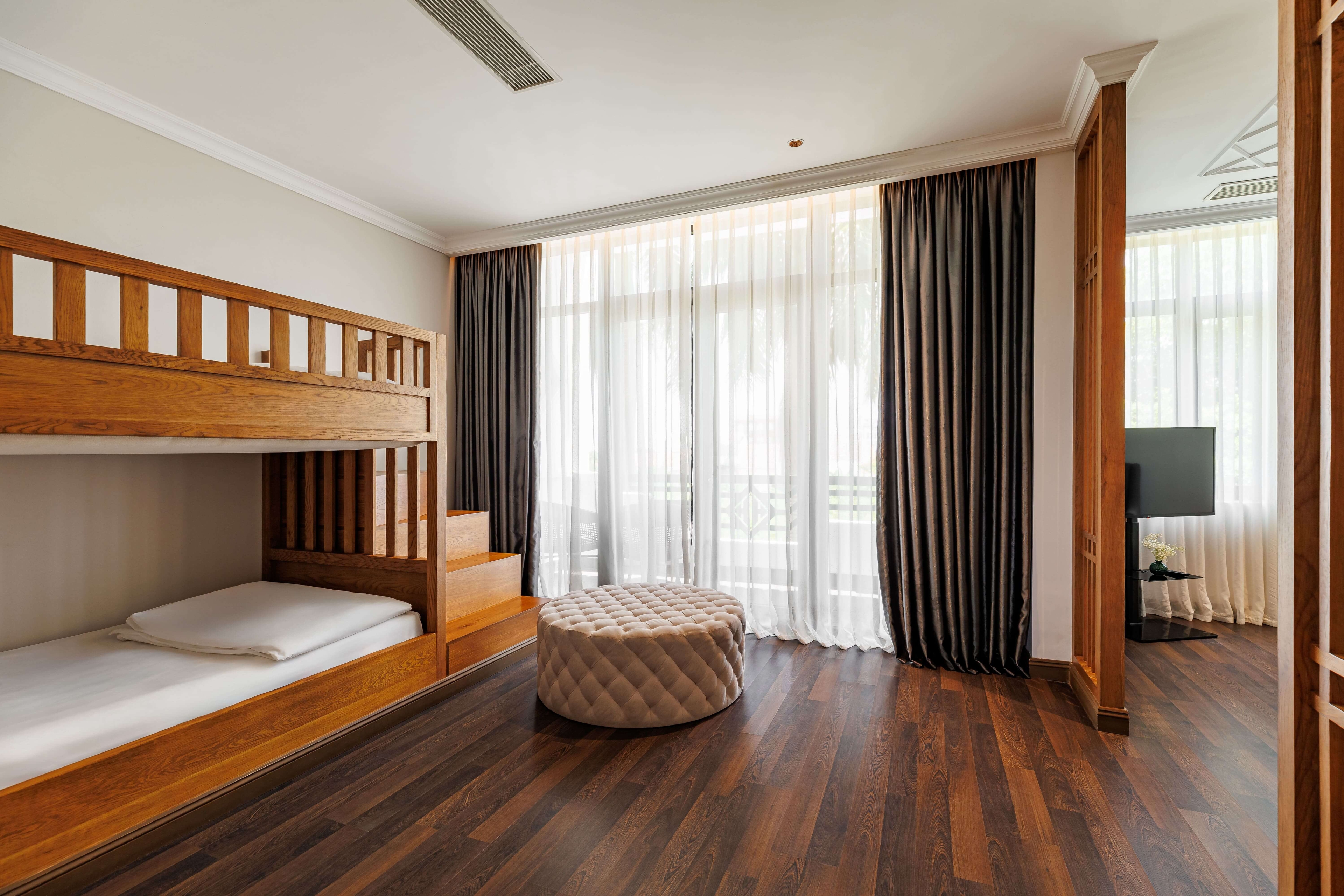 Bright bedroom featuring a wooden bunk bed with a lower sleeping area, accompanied by a round plush ottoman. Large windows with sheer curtains allow natural light to fill the space, creating a cheerful atmosphere. A television is visible in the corner, enhancing the room's functionality