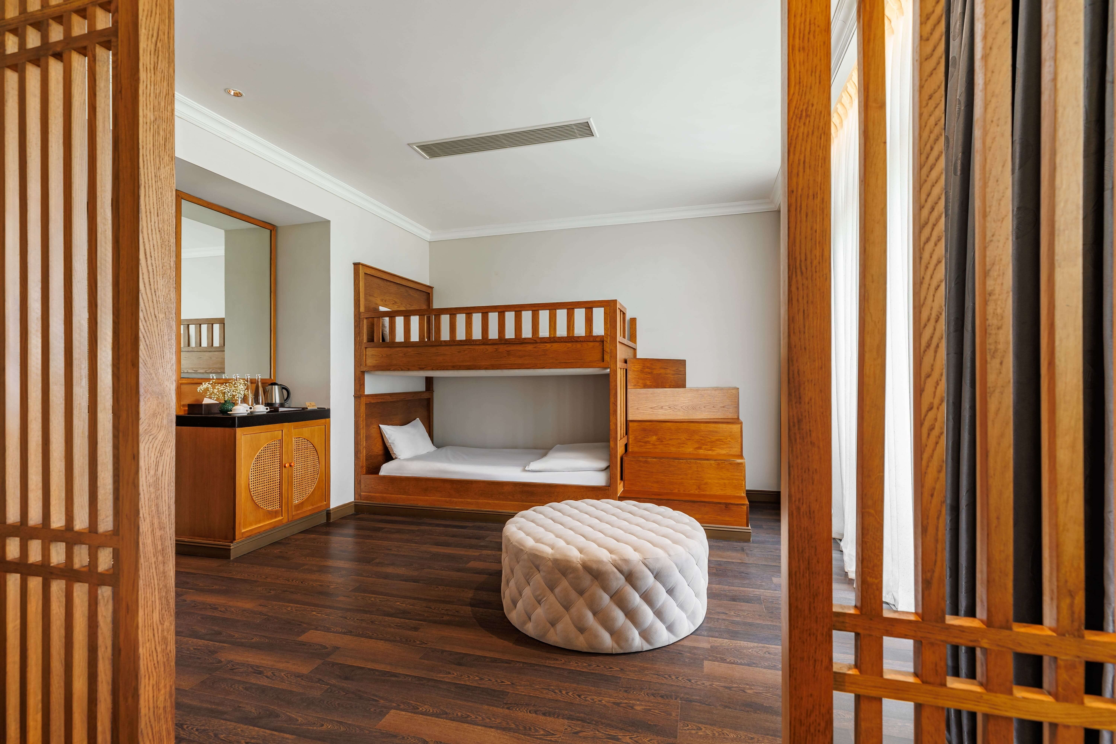 Cozy bedroom featuring a wooden bunk bed with a lower sleeping area and a plush round ottoman nearby. The room has warm wooden flooring and is partially enclosed by decorative wooden screens. A small kitchenette area is also visible in the background, adding functionality to the space