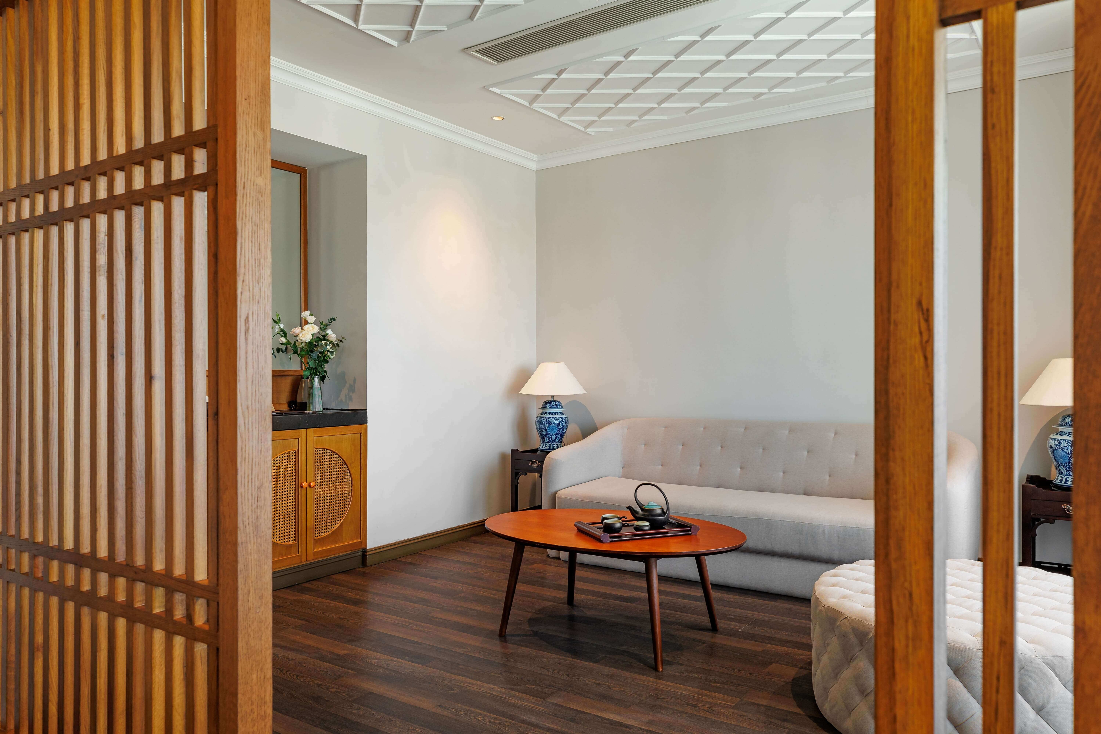 Charming living area with a light-colored sofa and a round wooden coffee table at the center. Soft lighting from two lamps creates a warm atmosphere. The room is partially separated by decorative wooden screens, enhancing the cozy and inviting décor