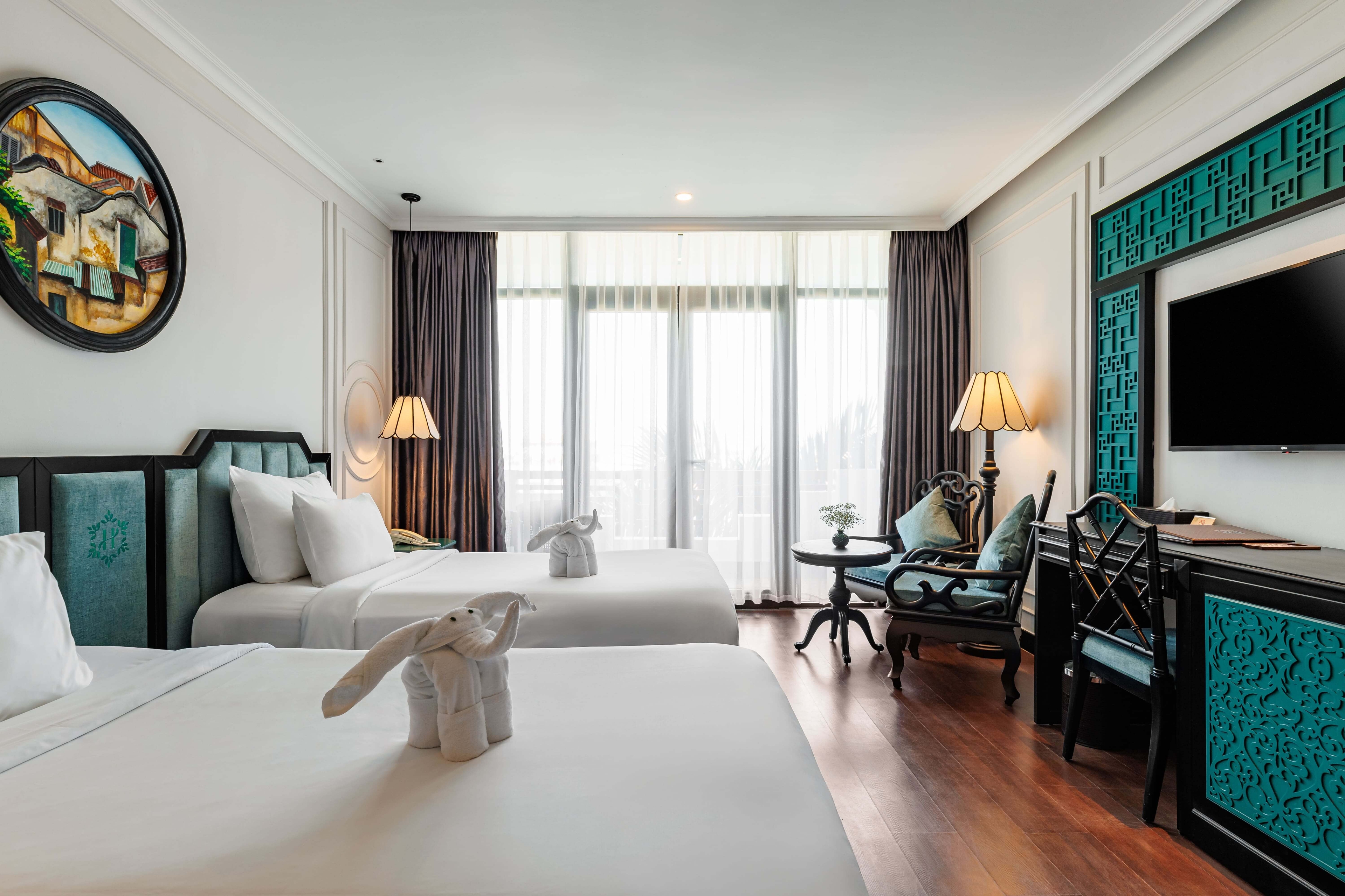 Bright hotel room with two twin beds, elegantly decorated with white linens and colorful accents. Large windows allow natural light, and the room features a small seating area with a table and a decorative TV unit