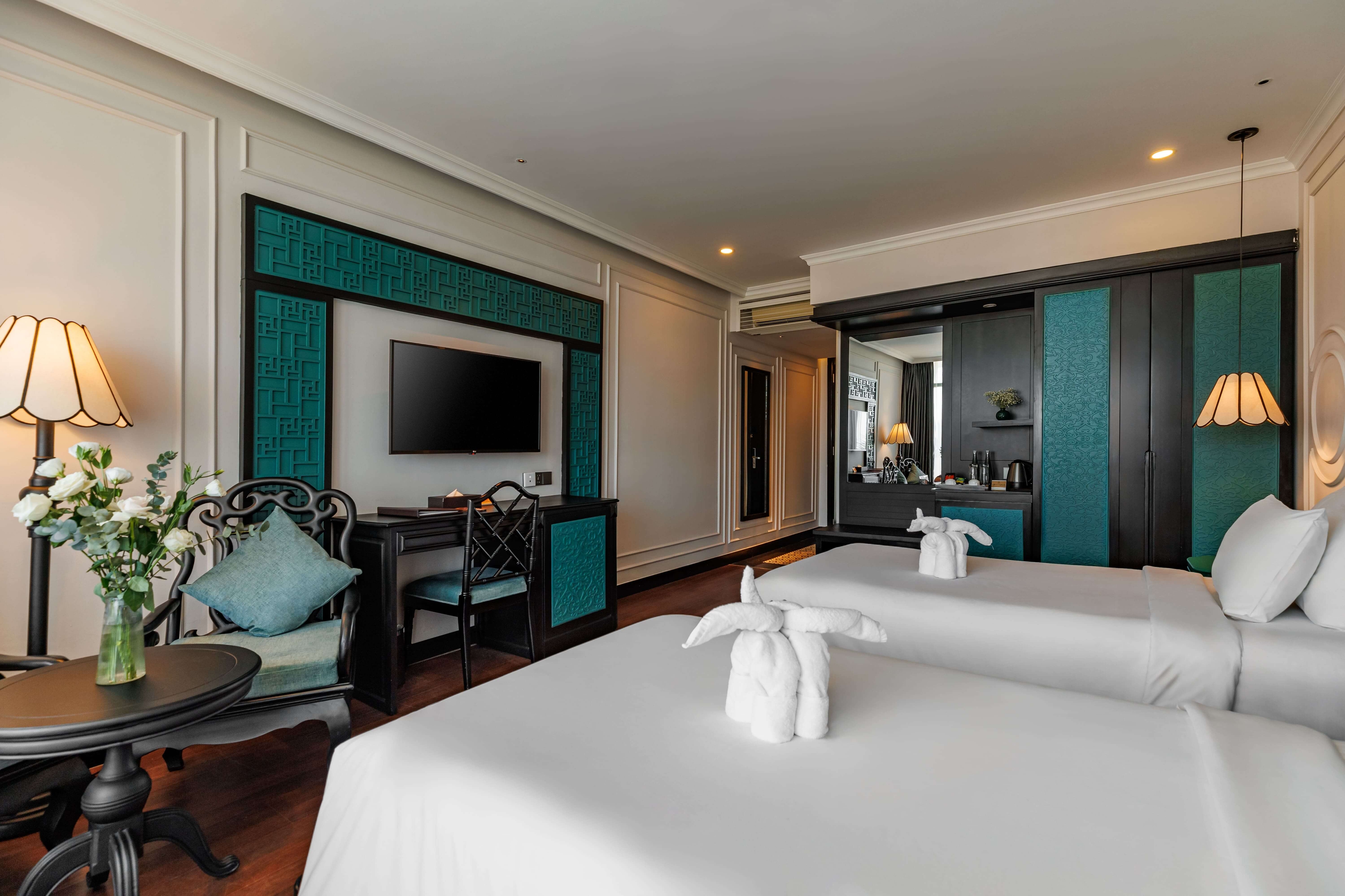 Stylish hotel room featuring two twin beds with white linens and decorative towels. The room includes a seating area with a small table, a TV mounted on the wall, and vibrant teal accents in the decor. Natural light filters in through large windows