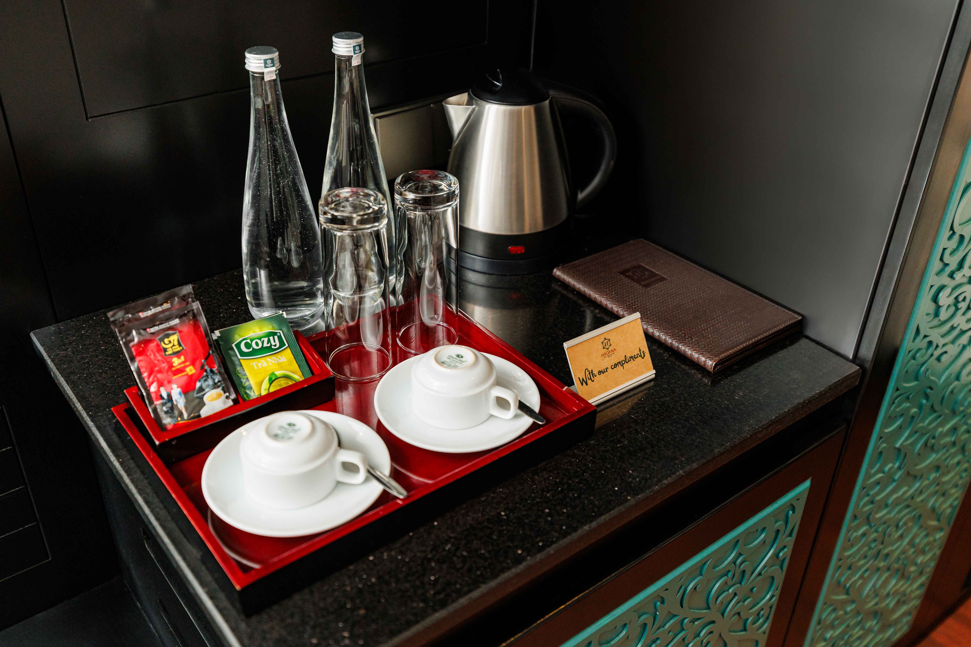 Hotel room amenities featuring two cups, a kettle, glass bottles, and a selection of tea bags on a decorative tray