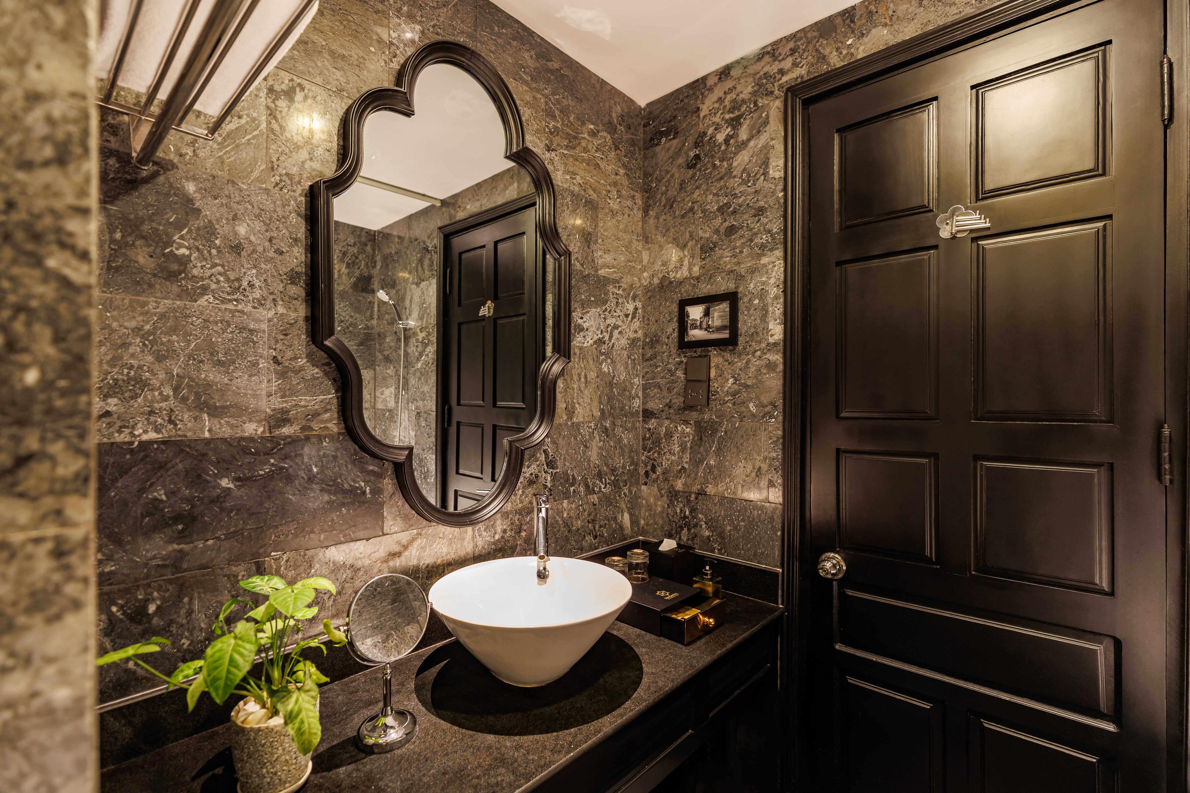 Sophisticated bathroom featuring dark marble walls and a stylish sink on a black countertop. A decorative mirror with an elegant frame hangs above the sink, and a small plant adds a touch of greenery. The door is a dark wood style, complementing the overall elegant decor