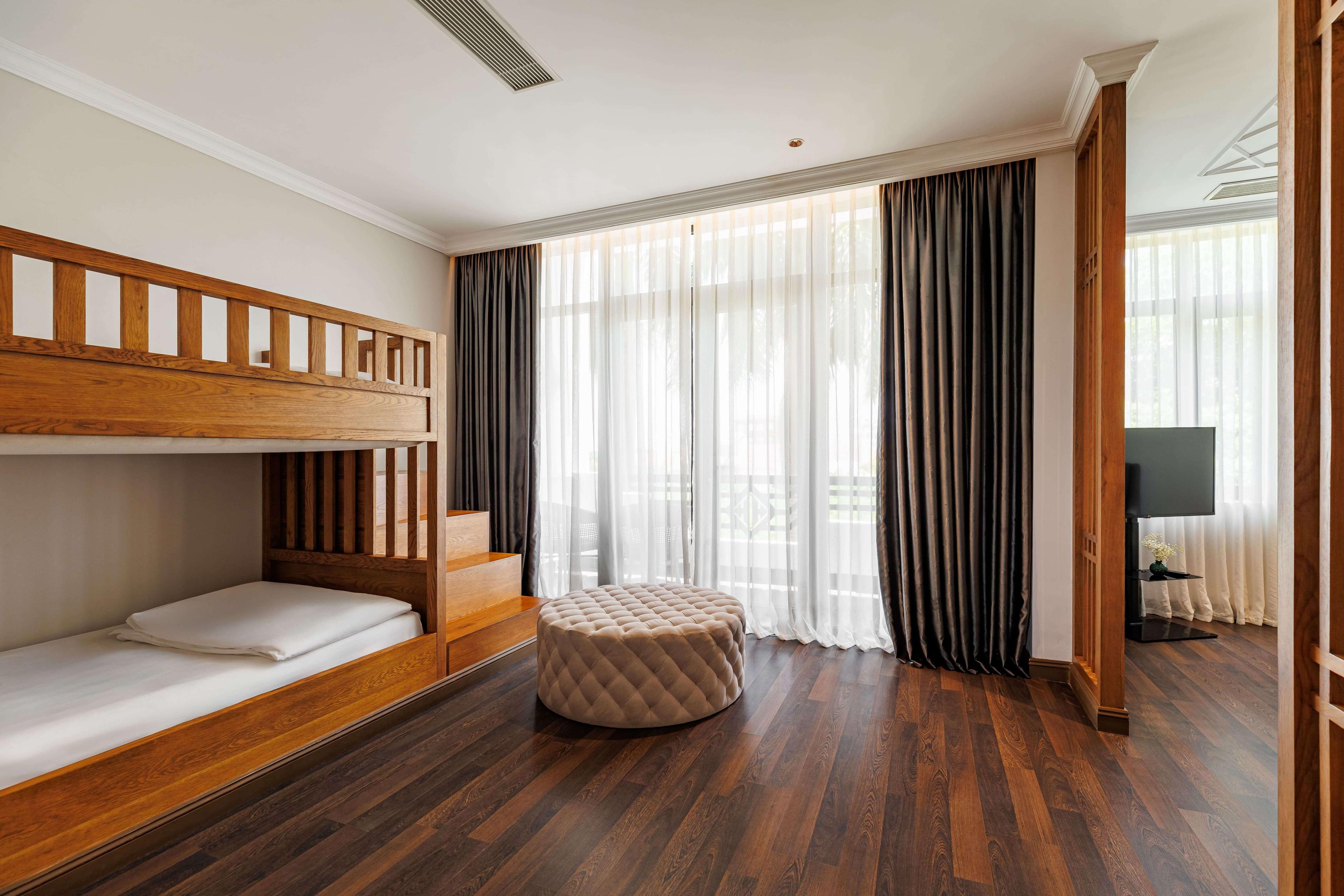 Bright bedroom featuring a wooden bunk bed with a lower sleeping area, accompanied by a round plush ottoman. Large windows with sheer curtains allow natural light to fill the space, creating a cheerful atmosphere. A television is visible in the corner, enhancing the room's functionality