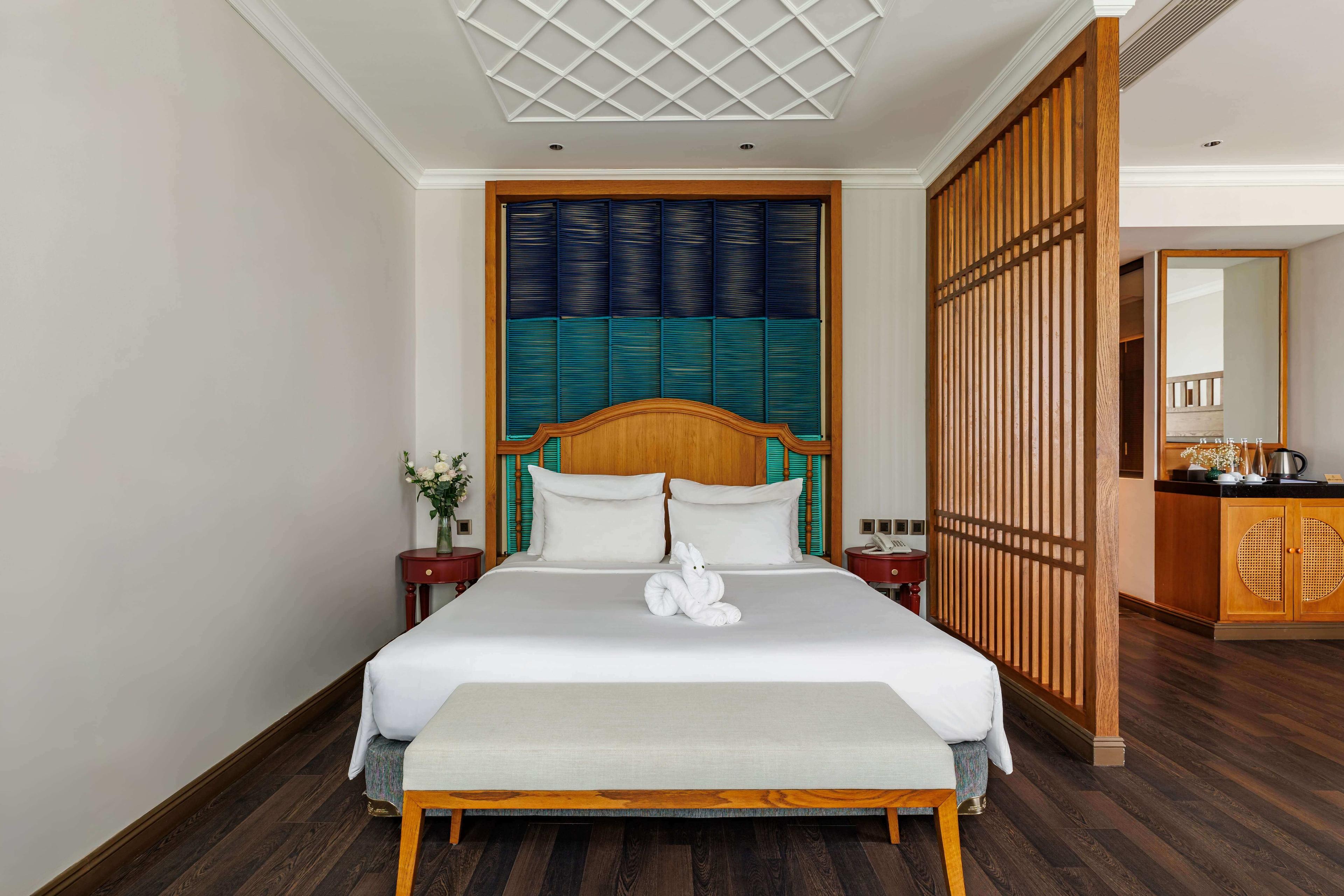 Bright and inviting bedroom featuring a queen-sized bed with crisp white linens and a decorative headboard in rich colors. A bench sits at the foot of the bed, and soft natural light fills the room. Partially visible wooden screens lead to a connecting space, enhancing the room's open feel